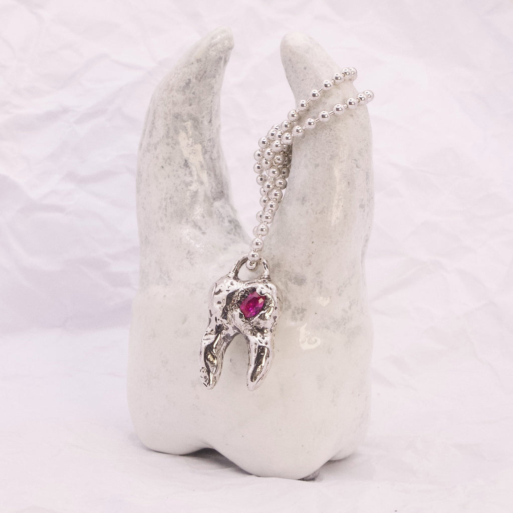 A silver-toned pendant necklace with a small winged figure and a red heart-shaped gemstone, displayed on a white sculptural stand against a soft white background.