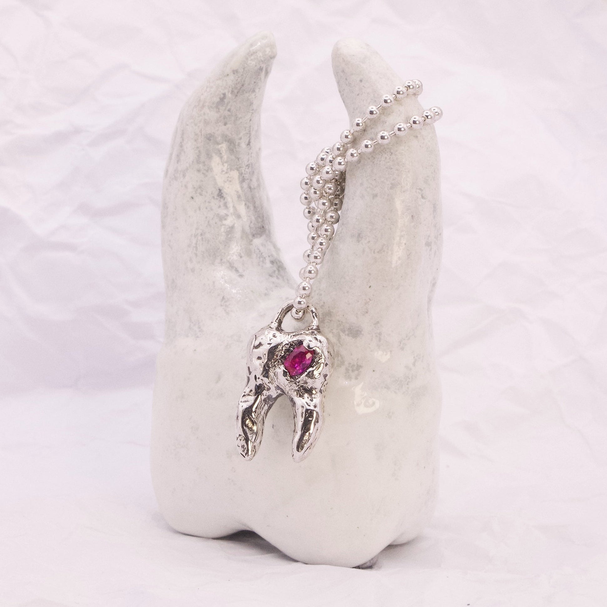 A silver-toned pendant necklace with a small winged figure and a red heart-shaped gemstone, displayed on a white sculptural stand against a soft white background.