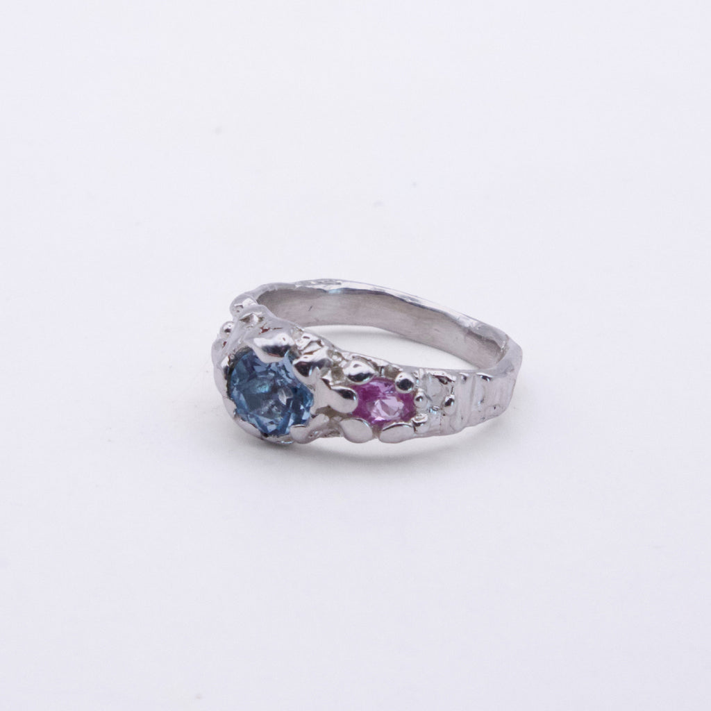 A silver ring featuring a round blue gemstone and a smaller round pink gemstone, set in an ornate, textured band on a plain light background.