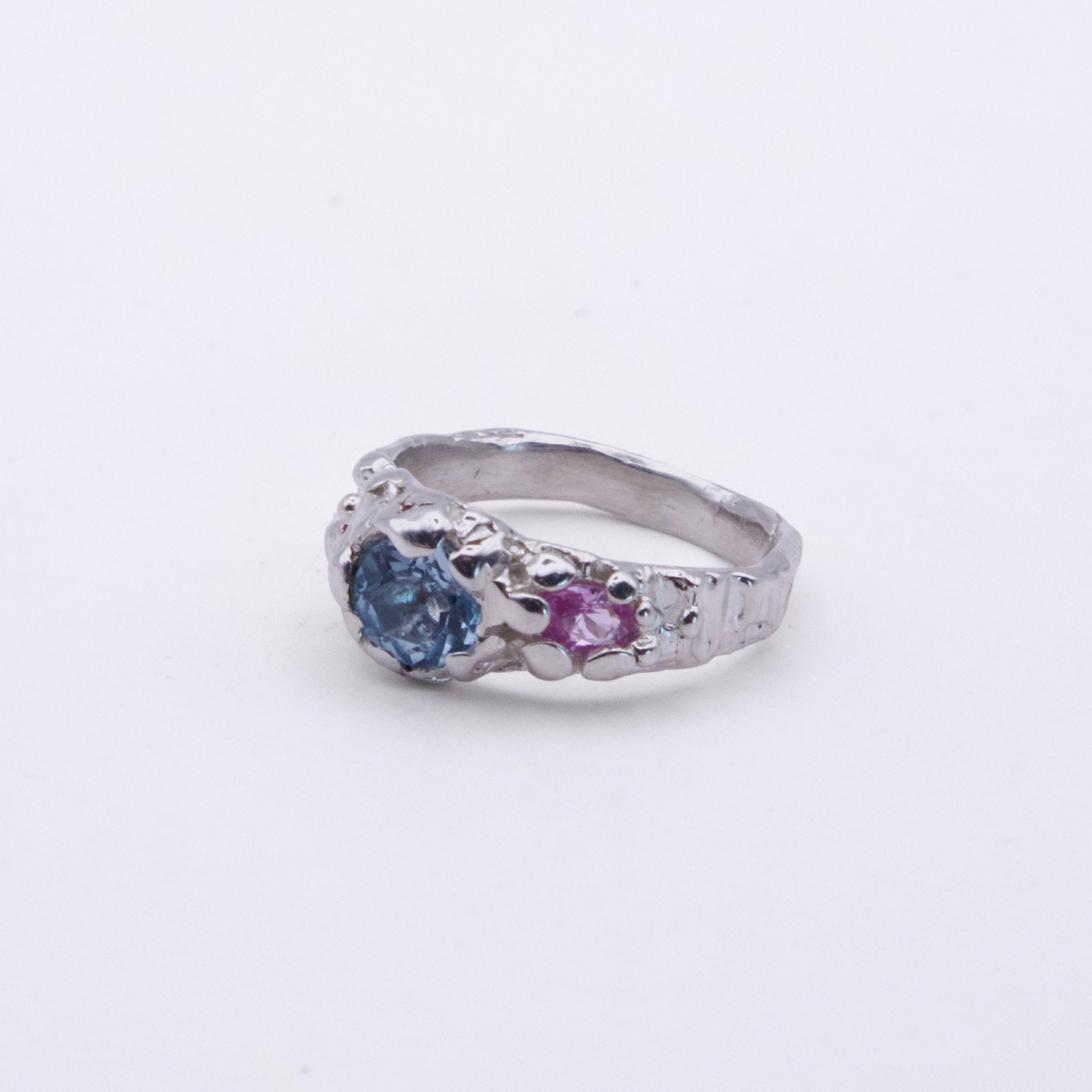A silver ring featuring a round blue gemstone and a smaller round pink gemstone, set in an ornate, textured band on a plain light background.