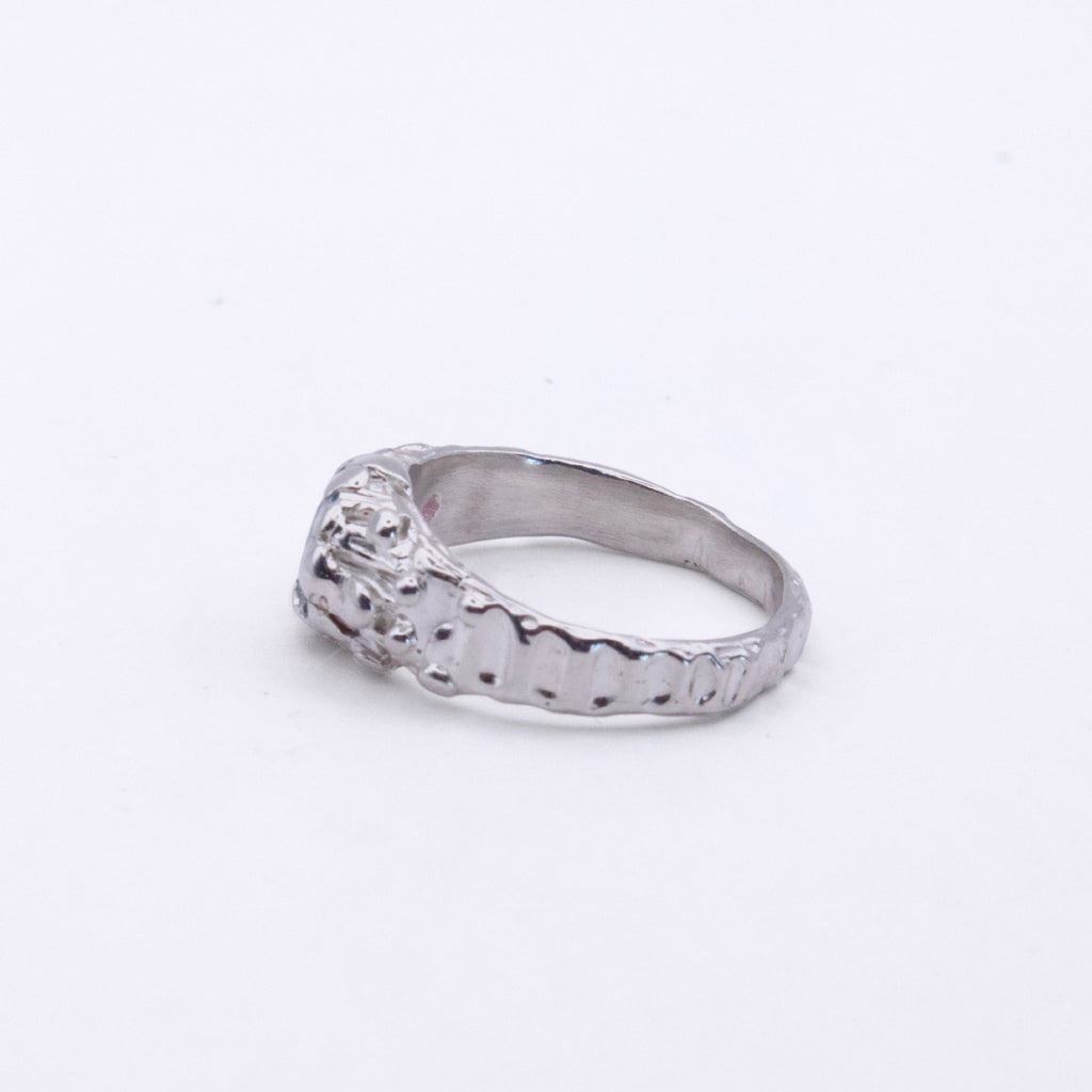 A silver ring with a textured, hammered band and a decorative, raised top section, photographed on a plain white background.