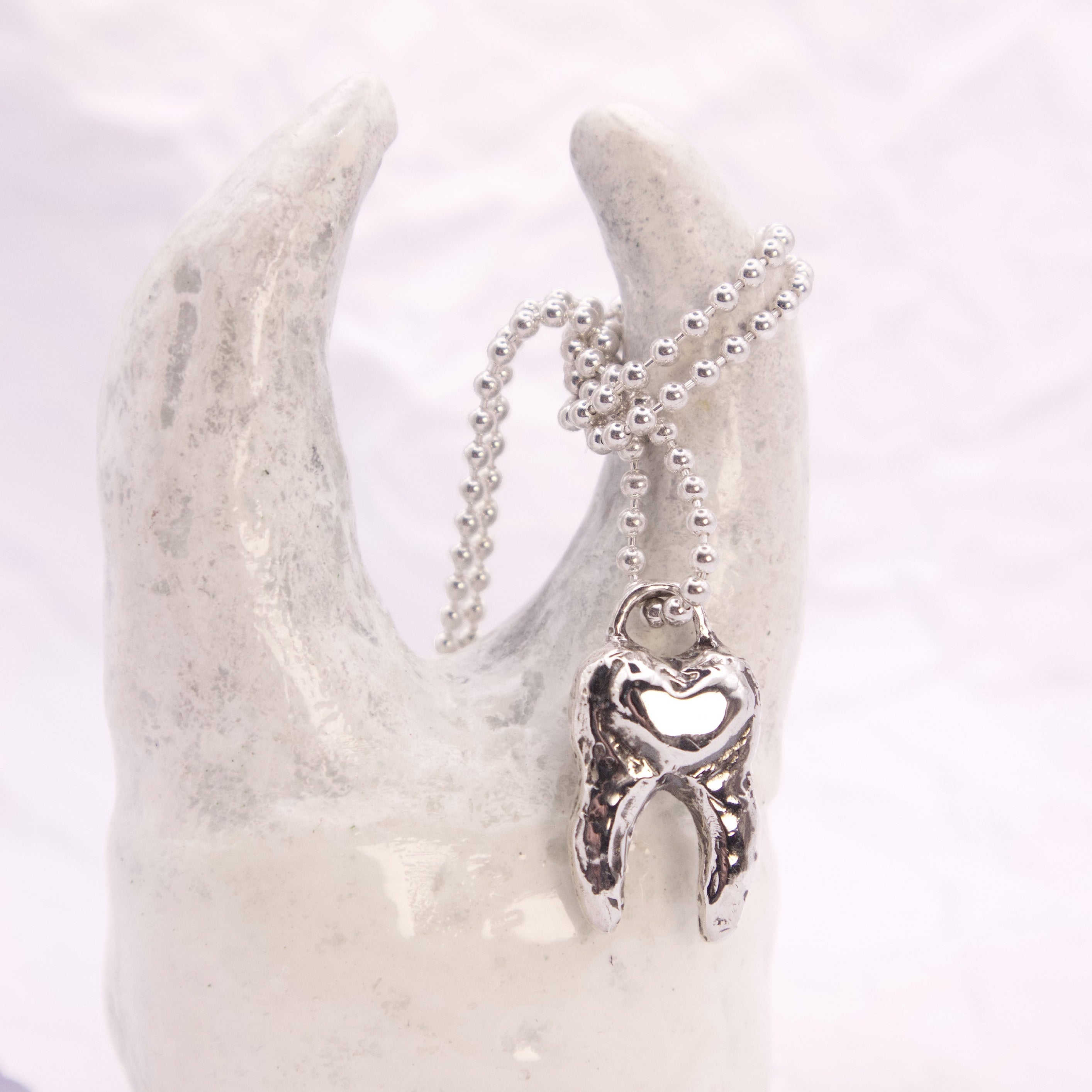 A silver tooth pendant necklace with a heart-shaped center, hanging from a silver ball chain and displayed on a white ceramic tooth-shaped stand.