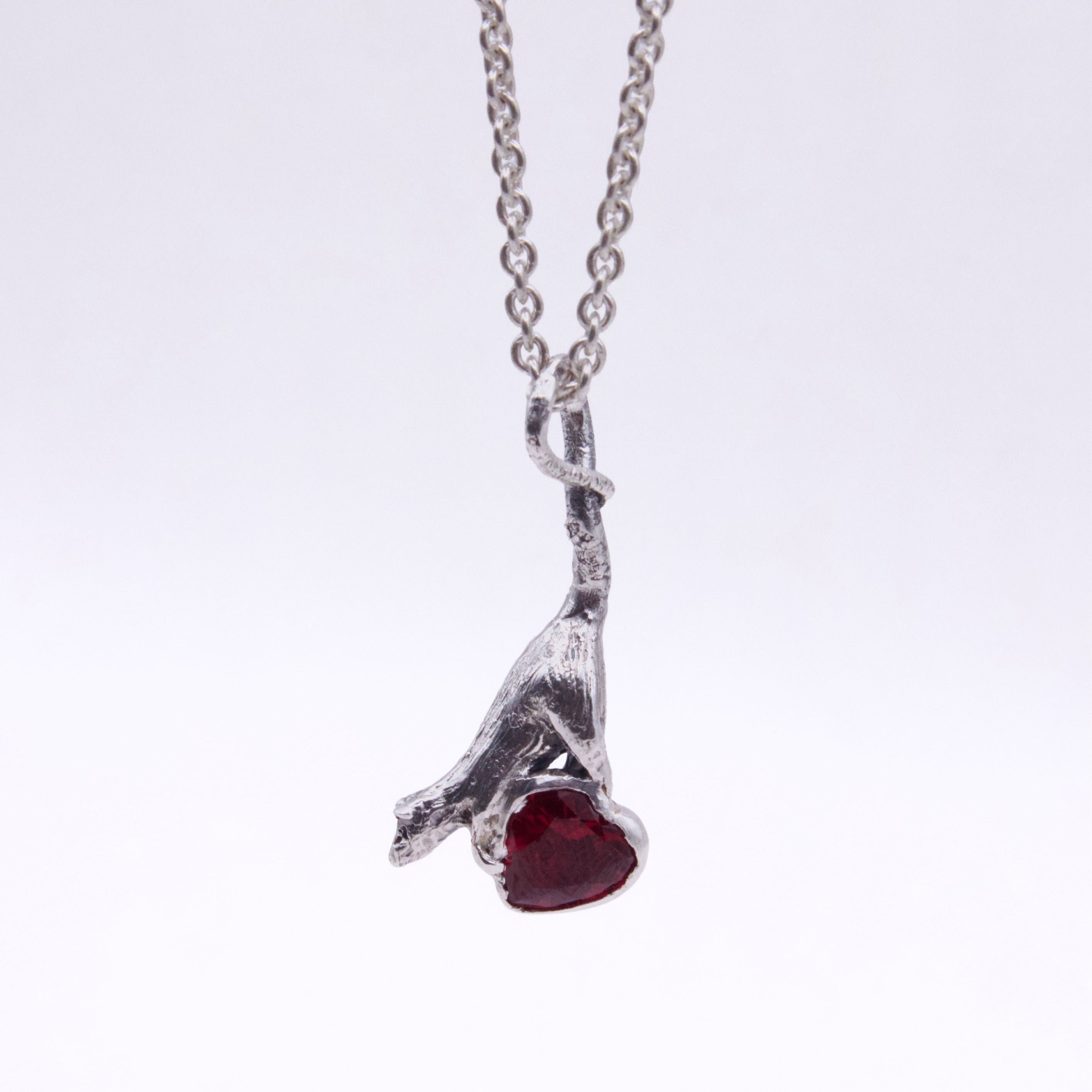 A silver-toned pendant necklace featuring a stylized rat with a heart-shaped dark red gemstone, suspended from a delicate chain against a plain white background.