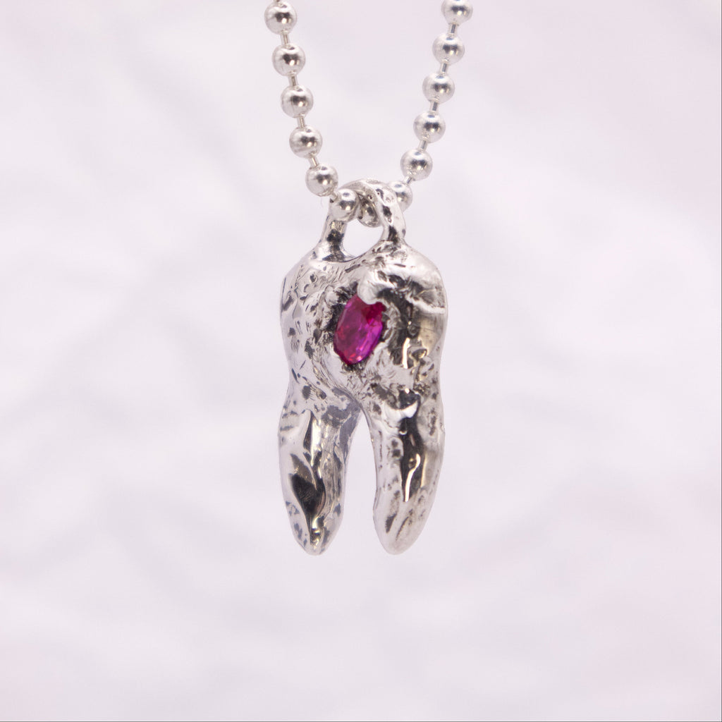 A silver ball-chain necklace with a sculpted tooth pendant featuring a small pink gemstone, shown against a soft white background.