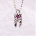 A silver ball-chain necklace with a sculpted tooth pendant featuring a small pink gemstone, shown against a soft white background.