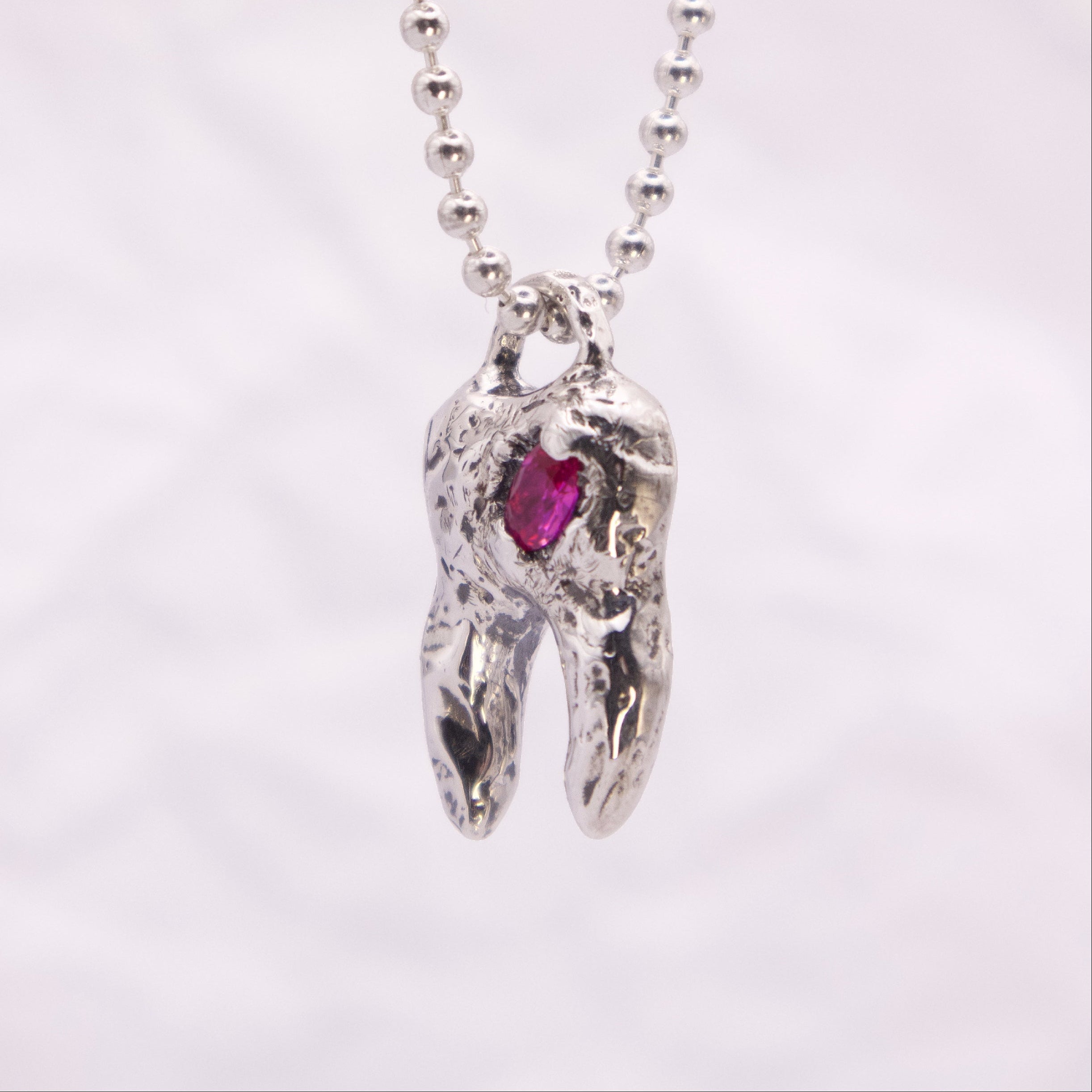 A silver ball-chain necklace with a sculpted tooth pendant featuring a small pink gemstone, shown against a soft white background.