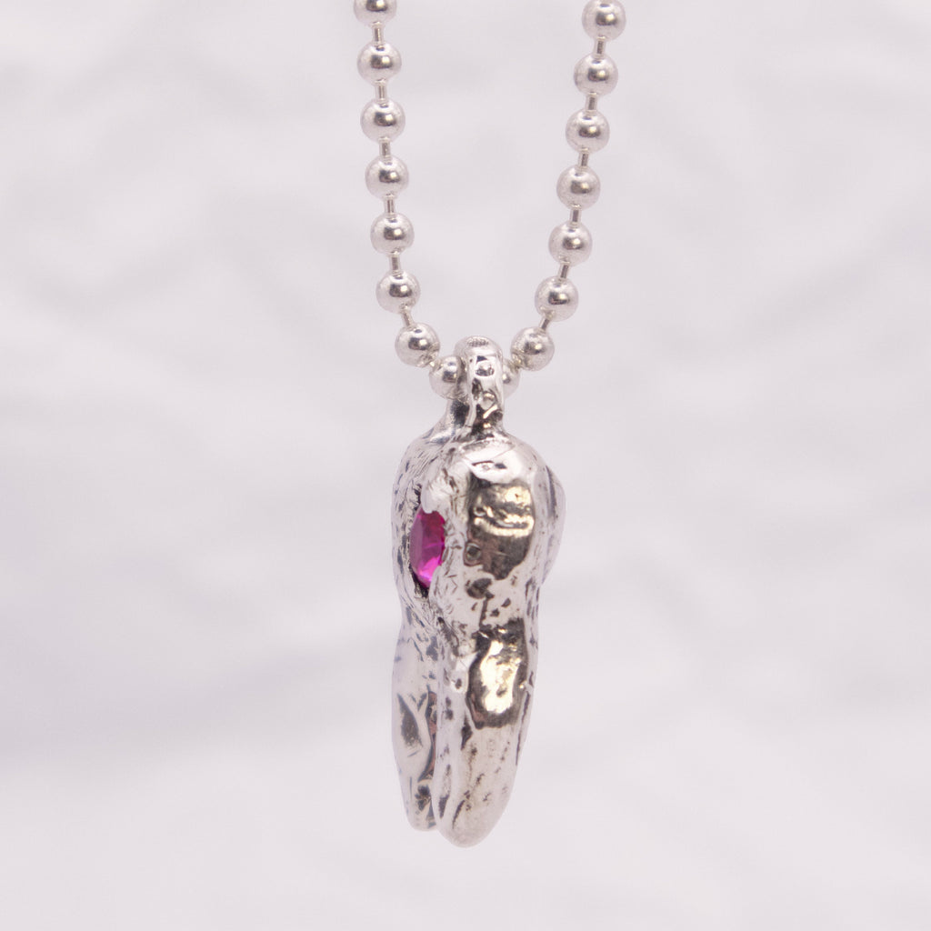 A silver beaded necklace with a textured, organic-shaped silver pendant featuring a small pink gemstone accent, shown against a light gray background.