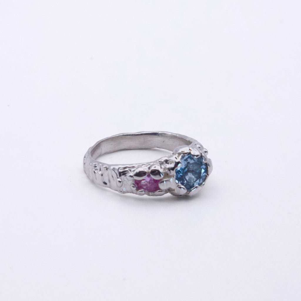 A silver ring featuring a round blue gemstone on one side and a smaller round pink gemstone on the other, set against a plain white background.
