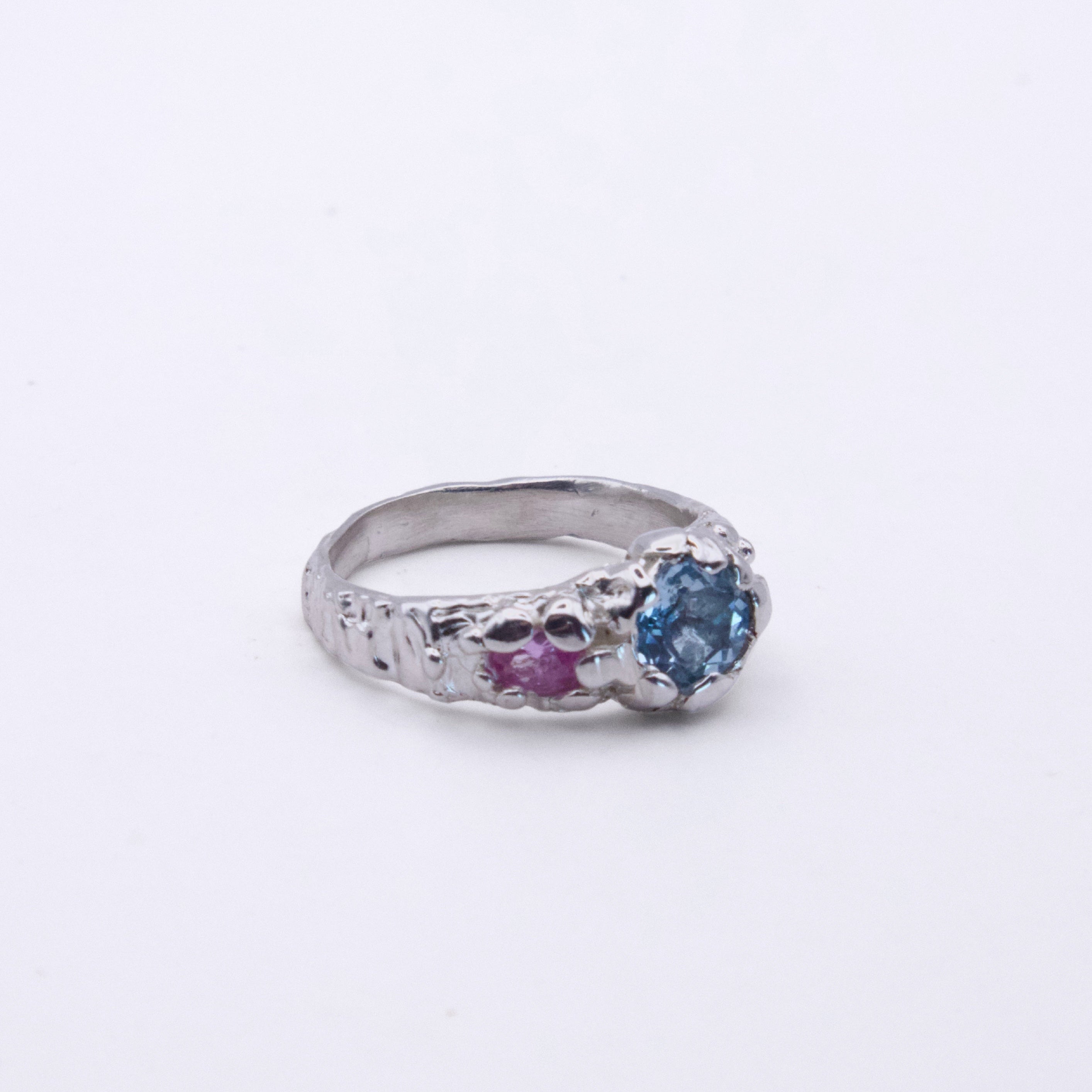 A silver ring featuring a round blue gemstone on one side and a smaller round pink gemstone on the other, set against a plain white background.