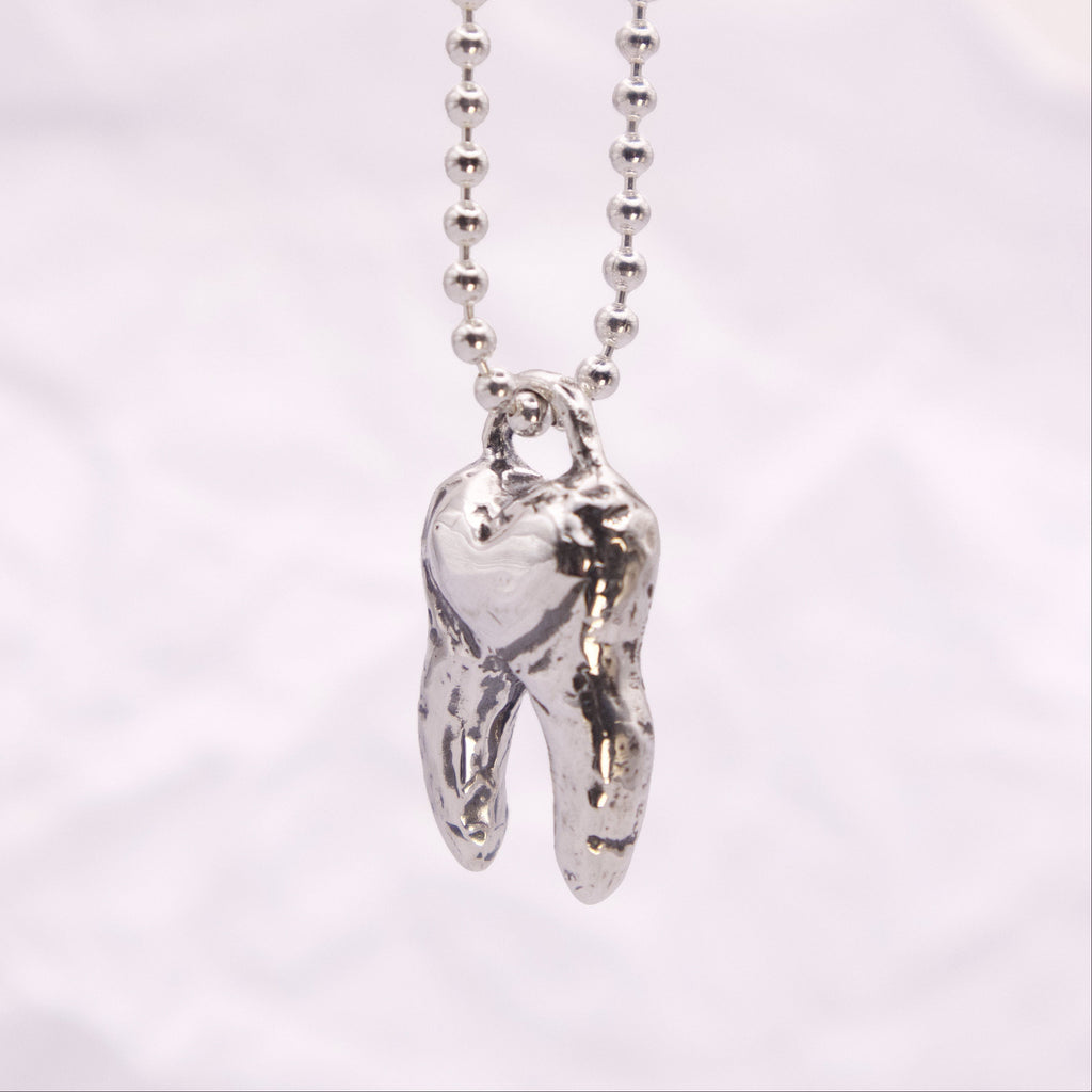 A silver ball chain necklace with a polished, textured silver molar tooth pendant, shown against a soft, light gray fabric background.