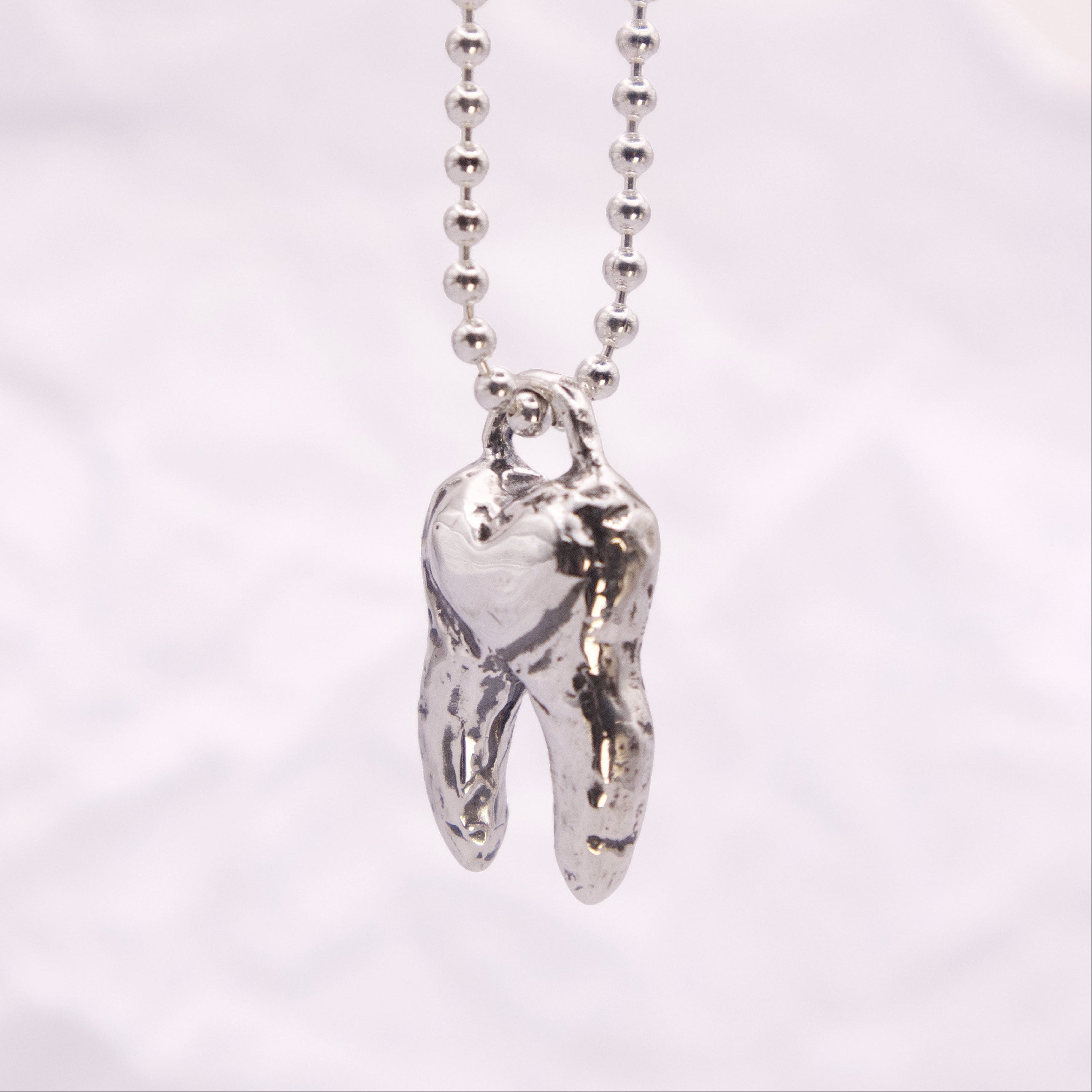 A silver ball chain necklace with a polished, textured silver molar tooth pendant, shown against a soft, light gray fabric background.