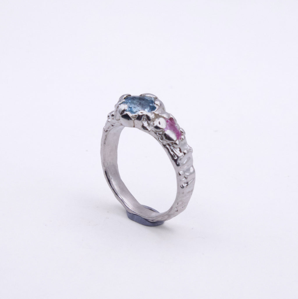 A silver ring featuring a textured, organic-shaped setting with a light blue oval gemstone and a small pink heart-shaped gemstone, set against a plain white background.