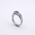 A silver ring featuring a textured, organic-shaped setting with a light blue oval gemstone and a small pink heart-shaped gemstone, set against a plain white background.