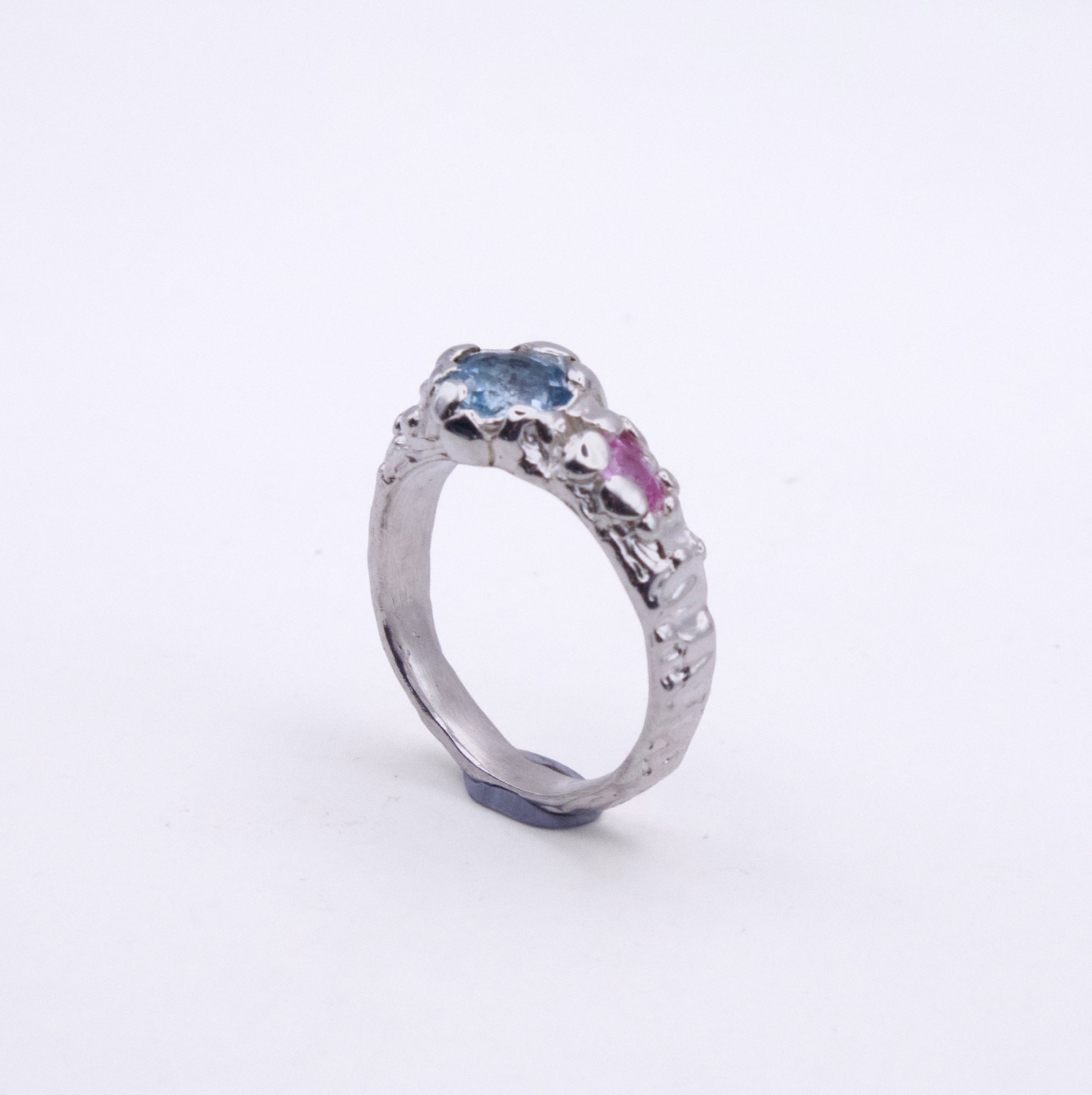 A silver ring featuring a textured, organic-shaped setting with a light blue oval gemstone and a small pink heart-shaped gemstone, set against a plain white background.