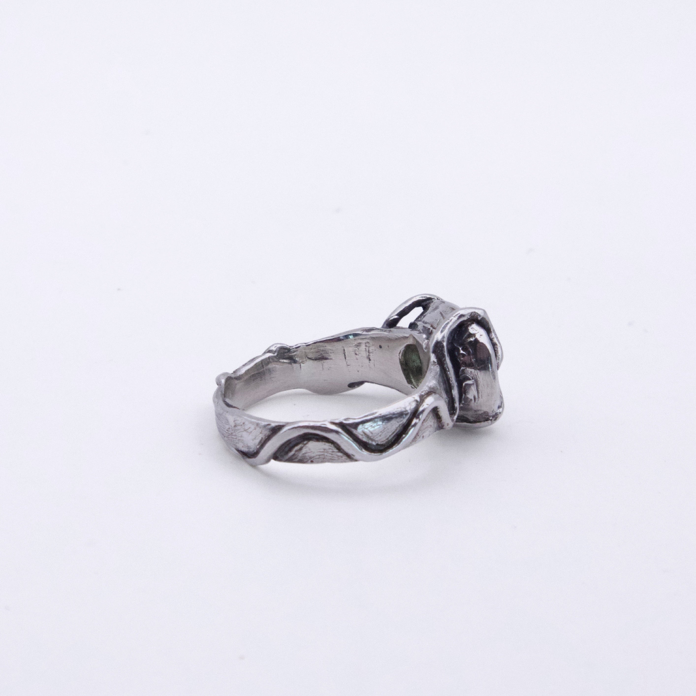 A silver-toned ring with a textured, organic wavy band and a dark, oxidized floral or knot-like setting, photographed on a plain white background.