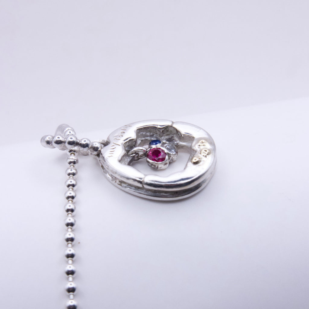 A silver Y2K pendant necklace with a small central pink gemstone and a blue accent stone, shown on a plain light gray background.