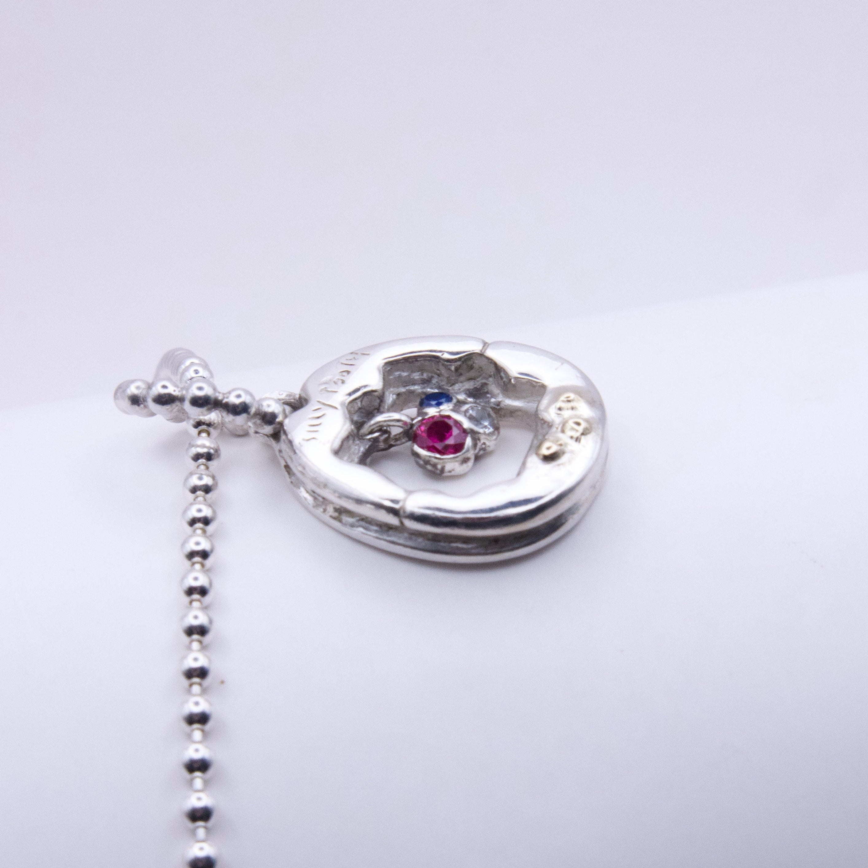 A silver Y2K pendant necklace with a small central pink gemstone and a blue accent stone, shown on a plain light gray background.